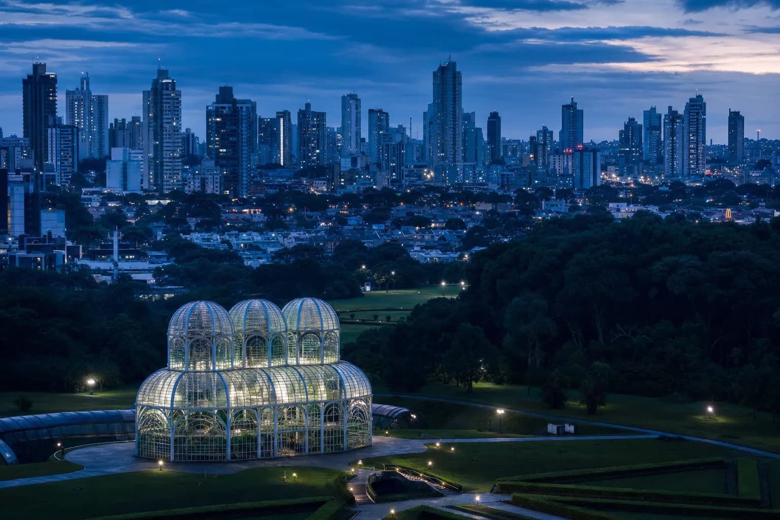 Estufa de vidro do Jardim Botânico de Curitiba com os prédios da cidade ao fundo, no fim de tarde